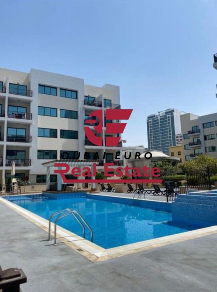 Modern apartment complex with white buildings surrounding a rectangular blue pool and poolside lounge chairs, overlaid with a red Dubai Euro Real Estate logo