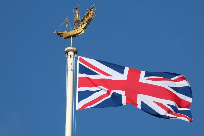 Union Jack flag waving on a flagpole with a gold ship weather vane against a blue sky.